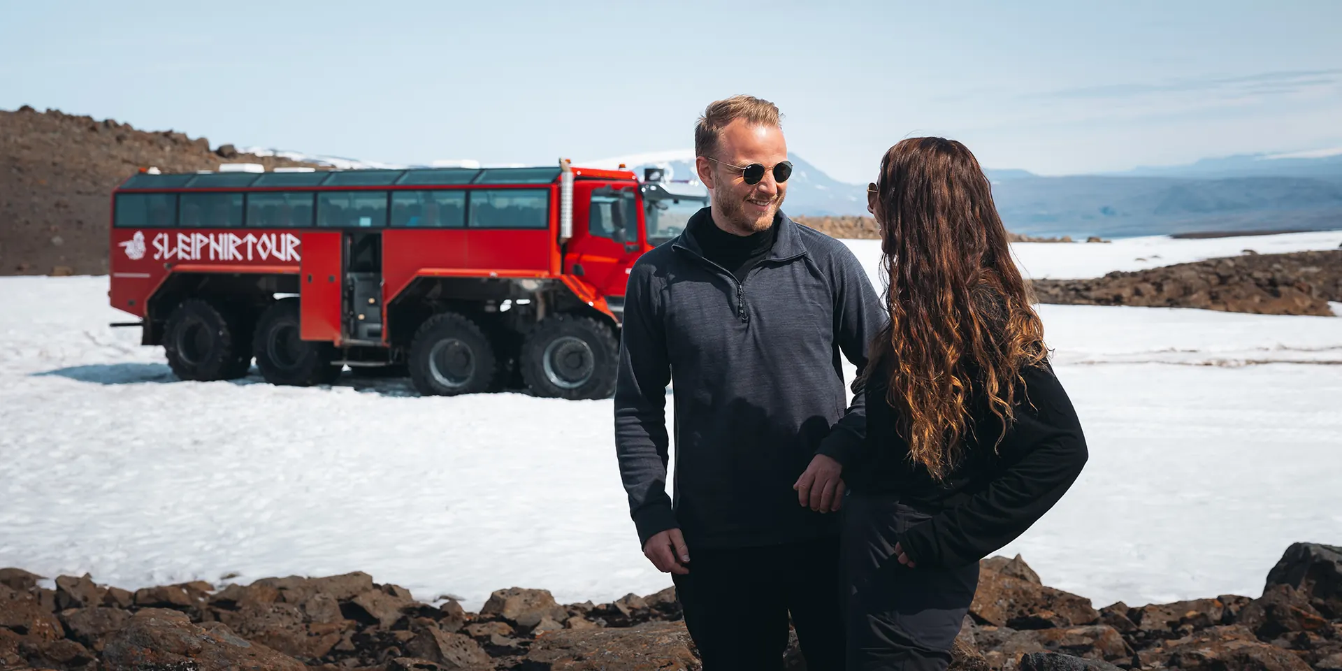 Two guests on the glacier with the red Sleipnir Monster Truck behind them